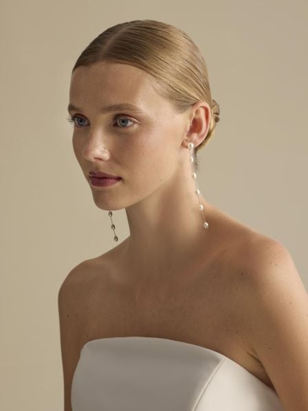 Woman with blonde hair wearing white strapless top and dangling pearl earrings against beige background