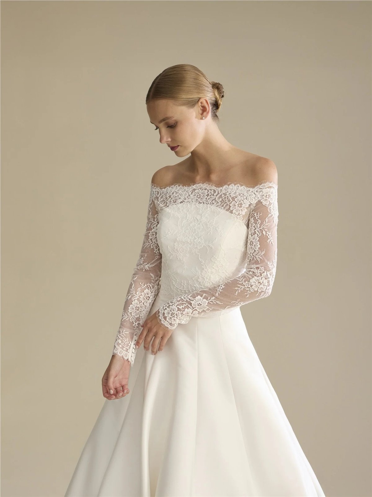 Woman in elegant off-shoulder white wedding dress with lace long sleeves and flowing skirt, looking down against neutral background