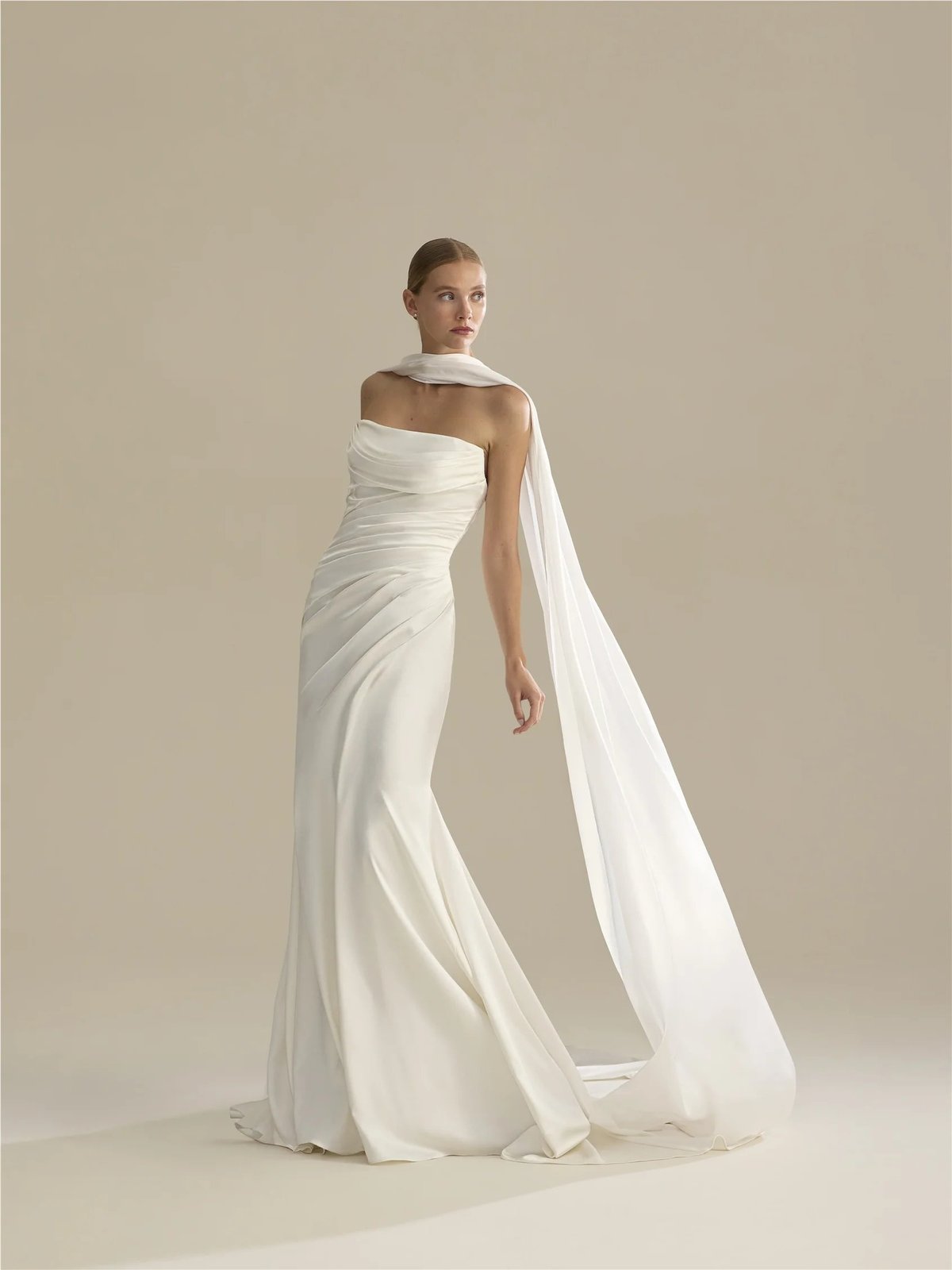 Woman wearing an elegant white draped gown with flowing cape against a neutral beige background