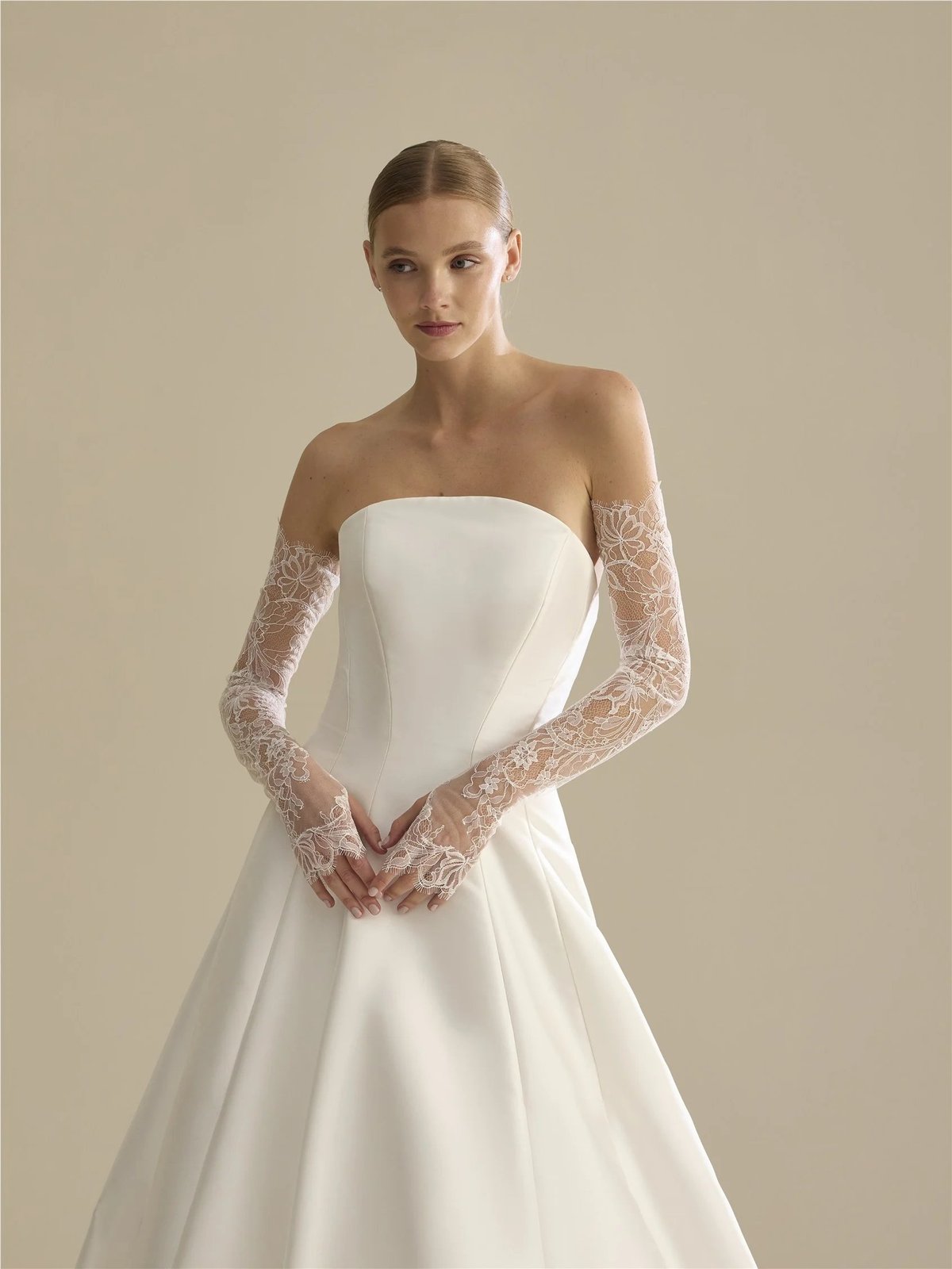 Woman wearing an elegant white strapless wedding dress with lace gloves against a beige background
