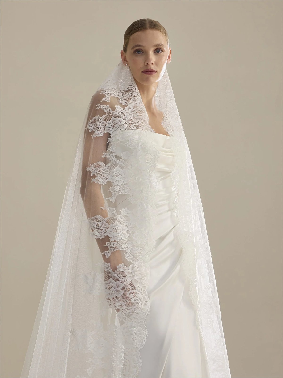 Woman in white lace wedding dress with draped lace veil against neutral background