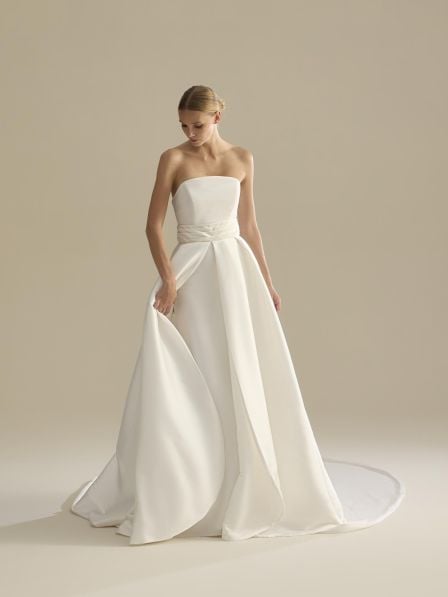 Woman wearing an elegant white strapless wedding gown with a flowing train and draped details against a neutral beige background